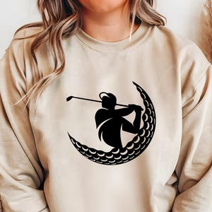 May include: A beige sweatshirt with a black silhouette of a golfer swinging a club inside a crescent moon shape made of golf balls.
