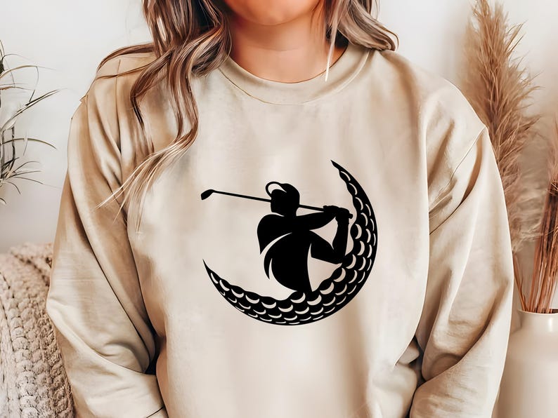 May include: A beige sweatshirt with a black silhouette of a golfer swinging a club inside a crescent moon shape made of golf balls.