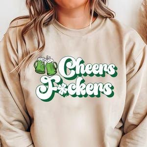 May include: Beige sweatshirt with the text "Cheers F*ckers" in white and green lettering. Two green beer mugs with white foam are on the left. A shamrock is below the word "F*ckers".