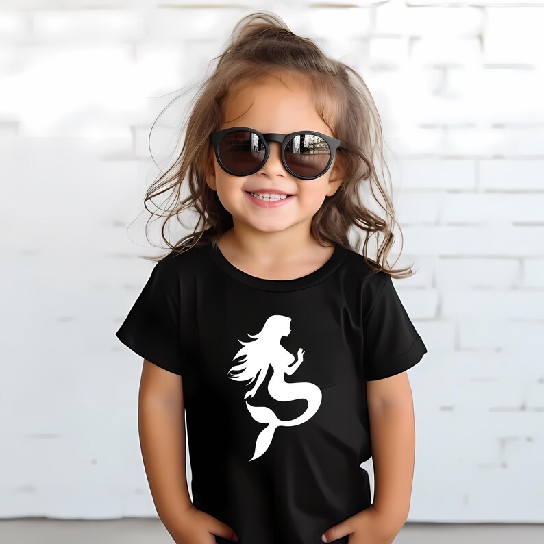 May include: A black t-shirt featuring a white mermaid graphic. The shirt is short-sleeved and appears to be made of a soft material. The image shows a young child wearing the shirt and sunglasses.