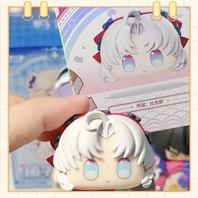 May include: A white-haired anime-style figurine with a red bow and blue accents. The figurine has large, colorful eyes and is held in front of a box with a matching image. The box has text in a foreign language.