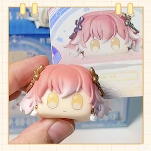 May include: A small, square-shaped figurine with a cartoon-like design. The figurine has pink hair, large yellow eyes, and gold-colored accessories. It is held in a person's hand, with a matching figurine visible in the background.