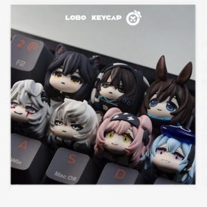 May include: A collection of anime-style keycaps in various colors, including black, white, pink, and blue, are arranged on a black keyboard. The keycaps feature detailed character designs with expressive faces. The text "LOBO KEYCAP" is visible.