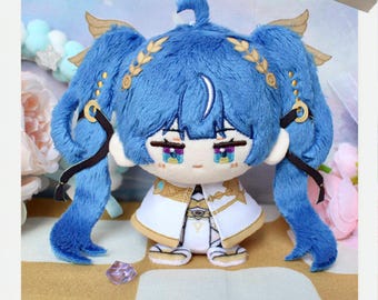 May include: A plush doll with blue hair styled in pigtails, adorned with gold accents. The doll has embroidered facial features, including large eyes and a small mouth. It wears a white and gold outfit. The image includes the words "MAKE" and "INSPIRATION".