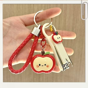 Puede incluir: Un juego de llaveros con una correa trenzada roja, un dije de manzana y un cortaúñas blanco. El dije de manzana tiene una cara sonriente y una hoja verde. Los llaveros son plateados.