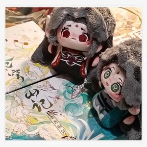 May include: Two plush dolls with detailed facial features and gray fur-like head coverings. One doll wears a black and red outfit, the other a teal and gray outfit. They rest on a surface with Asian-inspired artwork.