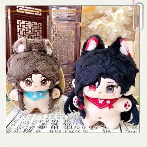 May include: Two plush dolls with anime-style features. One has brown hair and ears, a blue scarf, and a beige body. The other has black hair and cat ears with red accents, a red scarf, and a white body. Both dolls have detailed facial features.