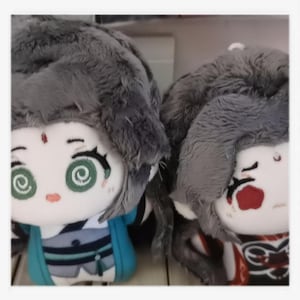 May include: Two plush dolls with detailed facial features and fluffy gray hair. One doll wears a teal and blue outfit, while the other has a red and black design. Both dolls have large, expressive eyes with spiral patterns.