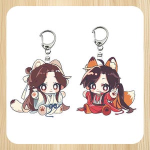 以下が含まれることがあります： 2つのアニメスタイルのキーホルダー。一体はクマ耳の白い衣装、もう一体はキツネ耳の赤い衣装。どちらも大きな目を持ち、シルバーのキーリングが付いています。