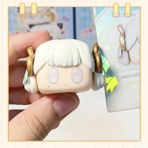 May include: A small, square-shaped figurine with a cartoonish face. The figurine has white hair, gold accents, and large, oval eyes. It is held by a hand, and a colorful background is partially visible.