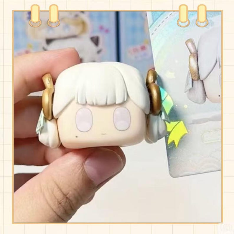 May include: A small, square-shaped figurine with a cartoonish face. The figurine has white hair, gold accents, and large, oval eyes. It is held by a hand, and a colorful background is partially visible.