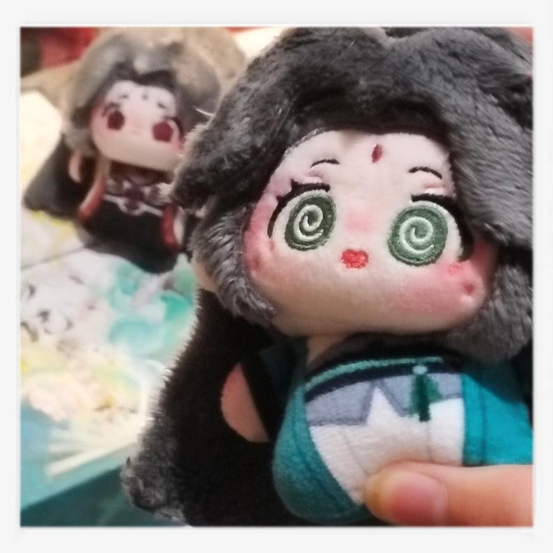 May include: Close-up of two plush dolls with detailed facial features. The dolls have large, spiral-shaped green eyes, rosy cheeks, and small red lips. They are dressed in teal and black outfits with gray hair. One doll is in focus, the other is blurred in the background.