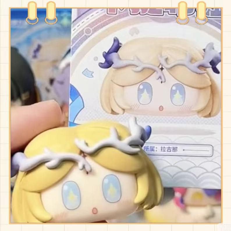 May include: A close-up of a figurine with a stylized face, featuring blonde hair and a white antler-like headpiece with purple accents. The eyes are large and blue, with a star-shaped design. The figurine is on a light-colored background.