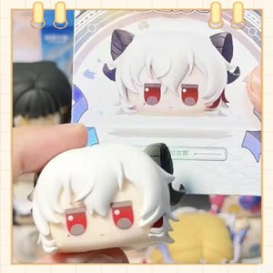 May include: A white figurine with a cartoon-like face, red eyes, and black horns. The figurine has white hair and is held in front of a matching illustration. The background is a light color with a decorative border.