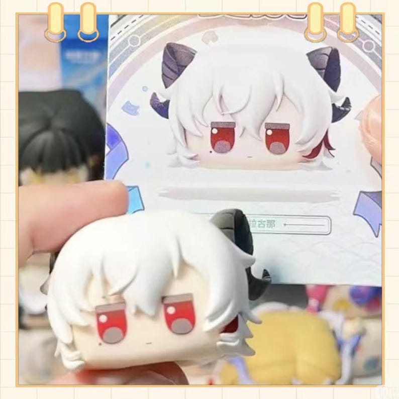 May include: A white figurine with a cartoon-like face, red eyes, and black horns. The figurine has white hair and is held in front of a matching illustration. The background is a light color with a decorative border.