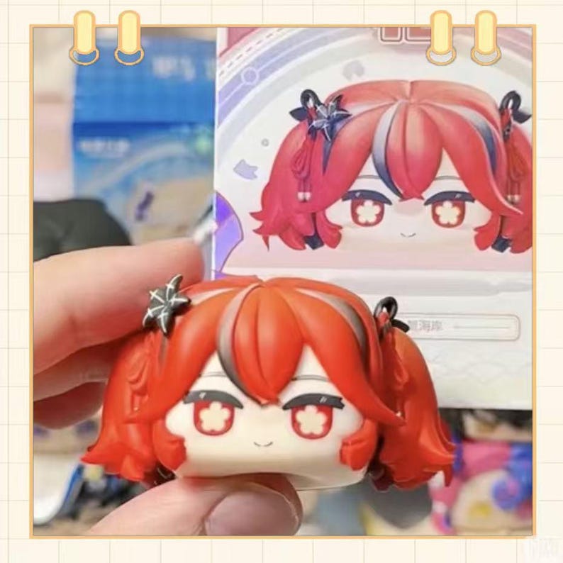 May include: A small, red-haired figurine with a smiling face and star-shaped hair accessories. The figurine has large, expressive eyes and is held in a person's hand. A second image of the figurine is visible in the background.