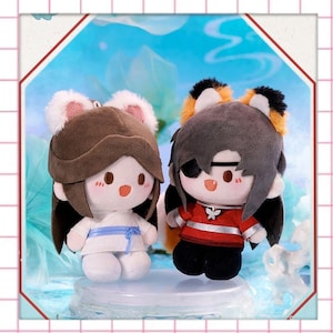 May include: Two plush dolls with anime-style features. One doll wears a brown hat with pink ears and a white outfit with a blue sash. The other doll has a black eye patch, a red and black outfit, and orange and black ear accents.