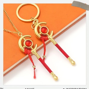 Op de afbeelding: Goudkleurige ketting met hanger en sleutelhanger in de vorm van een halve maan met rode accenten. De hanger en sleutelhanger hebben een rode edelsteen in het midden en een rode kwast. De sleutelhanger heeft een gouden ring.
