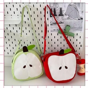 May include: Two apple-shaped crossbody bags. One is light green, the other is red. Both bags feature a white apple slice design with brown seed details and a green leaf. They have long straps for shoulder or crossbody wear.