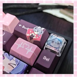 Puede incluir: Primer plano de un teclado mecánico con teclas personalizadas. Las teclas son de varios tonos de rosa y morado, con diseños de personajes de estilo anime. Algunas teclas tienen texto como "Back" y "Del".