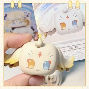 May include: A cream-colored, square-shaped figurine with yellow accents, featuring orange and blue eyes, and a small red star. The figurine has small wings and a curled top. The image also shows a blurred image of a similar figurine and text in a foreign language.