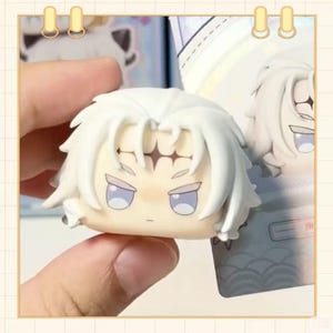 May include: A small, white figurine head with spiky hair and a stern expression. The figurine has light blue eyes and a neutral skin tone. The head is held in a hand, with a blurred background showing similar figurines.