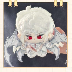 May include: A plush doll with white fur hair and red eyes, wearing a white outfit with a detailed design. The doll has gray wings and is held against a dark background.