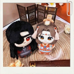 May include: Two plush dolls with detailed embroidered faces and outfits. One doll has black hair and a red eye patch, the other has brown hair and a decorative headpiece. The dolls are on a woven surface with a decorative screen in the background.
