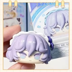 May include: A close-up of a small, collectible figurine with a stylized anime aesthetic. The figure has lavender hair, a white frilly cap, and a serene expression. The figure is held in a hand, with a portion of the packaging visible in the background.