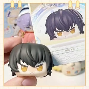 May include: A small, black-haired figurine with a stern expression and yellow eyes is held in a hand. The figurine is a miniature version of a character from a cartoon or anime. A card with a matching illustration is behind it.
