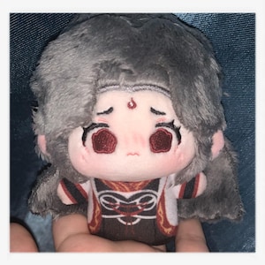 May include: A plush doll with a sad expression, featuring gray hair and a detailed outfit. The doll has a light pink face with red eyes and a small red mark on its forehead. The outfit is a mix of white, brown, and red.