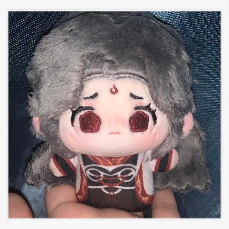 May include: A plush doll with a sad expression, featuring gray hair and a detailed outfit. The doll has a light pink face with red eyes and a small red mark on its forehead. The outfit is a mix of white, brown, and red.