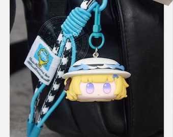 May include: A yellow-haired anime-style charm with a white hat and blue accents, attached to a blue and black lanyard. The lanyard has a blue clasp and a tag with text and a magnifying glass graphic. The charm is hanging against a black background.