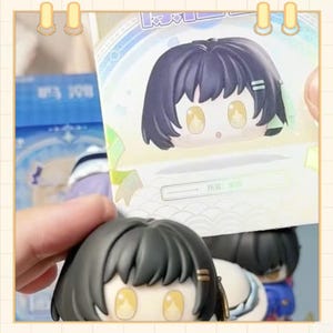 May include: A close-up of a small, cartoon-style figure with a large head and big eyes. The figure has black hair and is holding a card with a similar image. The figure is in a light-colored box.
