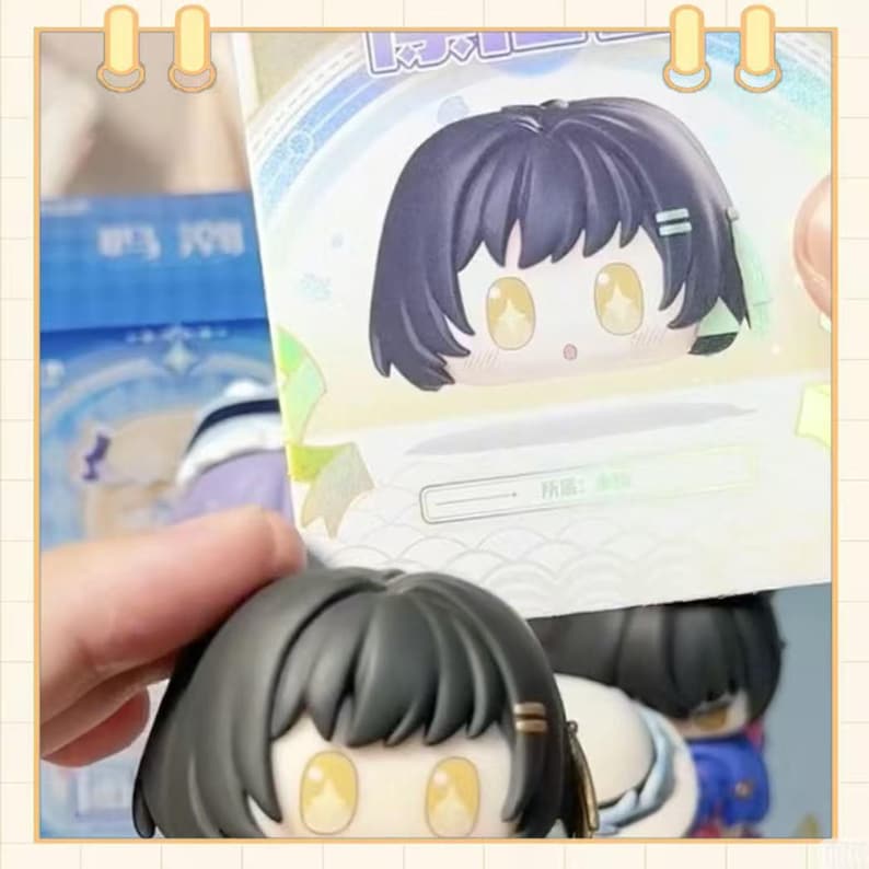 May include: A close-up of a small, cartoon-style figure with a large head and big eyes. The figure has black hair and is holding a card with a similar image. The figure is in a light-colored box.