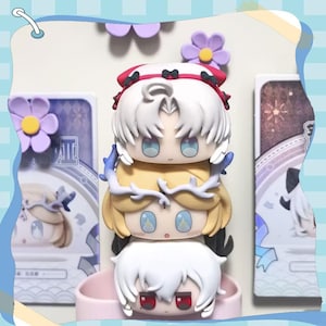 May include: A stack of four chibi-style figures with white, yellow, and red hair, and blue eyes. The figures are stacked on top of each other, with a pink base. The background includes decorative elements and a light blue border.