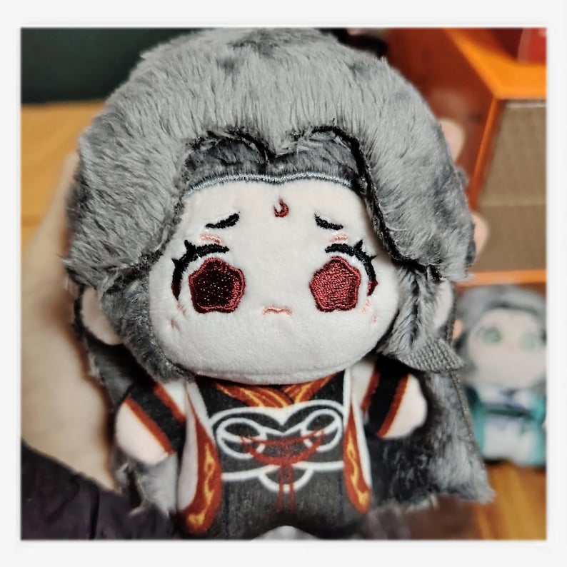 May include: A plush doll with a sad expression, featuring gray hair and a detailed outfit in black, red, and gold. The doll has embroidered facial features and is held in a person's hand.