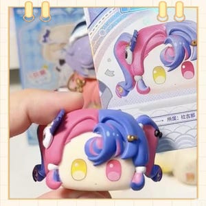 May include: A colorful collectible figurine with a split pink and blue hairstyle, yellow and pink eyes, and a cream-colored face. The figurine is part of a series, with a matching box in the background.