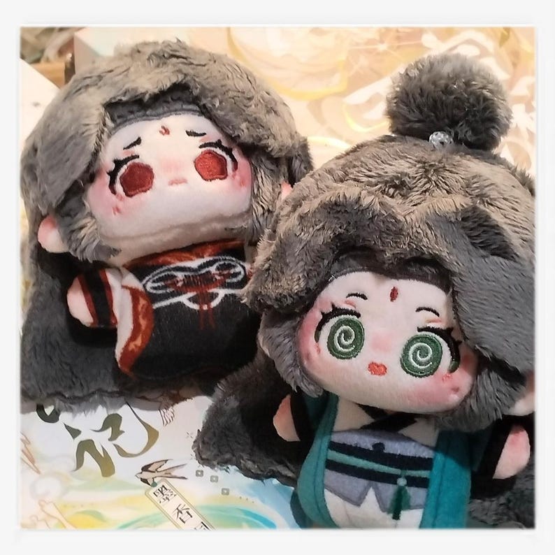 May include: Two plush dolls with detailed embroidered faces and gray fur hoods. One doll wears a black and red outfit, the other a teal and gray outfit. Both dolls have large, expressive eyes and are set against a patterned background.