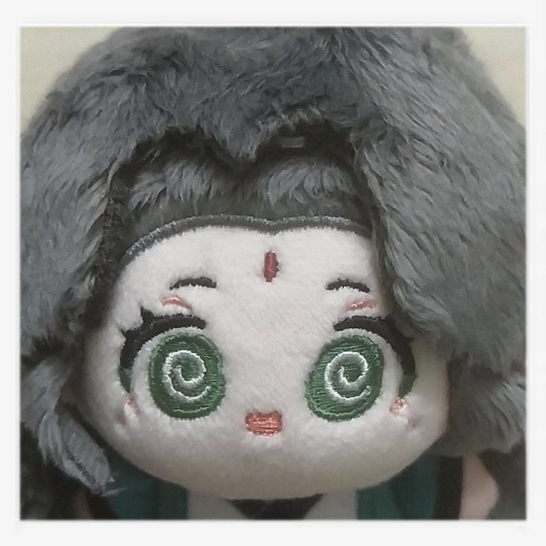 May include: A plush doll with a white face, green spiral eyes, and a small red mark on the forehead. The doll has gray, fluffy hair and a small, embroidered pink mouth. The doll is wearing a green and white outfit.