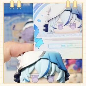 May include: A collectible figurine with a cartoon character design. The figure has a white and blue color scheme, with light blue hair and a matching hat. The figure is held in a person's hand, with a box in the background.