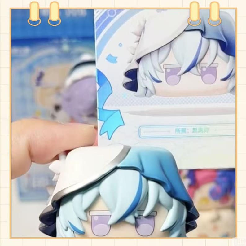 May include: A collectible figurine with a cartoon character design. The figure has a white and blue color scheme, with light blue hair and a matching hat. The figure is held in a person's hand, with a box in the background.