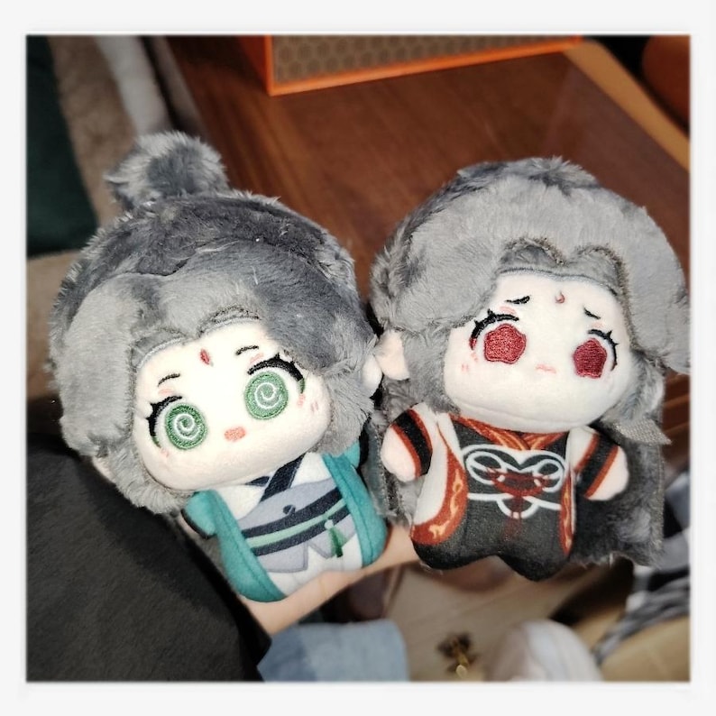 May include: Two plush dolls with gray hair and detailed facial features. One doll wears a teal and white outfit, the other a black and red outfit. Both dolls have a cartoonish design with large eyes.