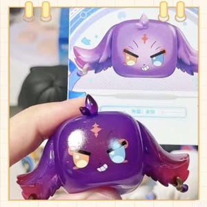 May include: A purple, cube-shaped figurine with large, floppy ears and a small horn. The figurine has two different colored eyes, one yellow and one blue, and a small, mischievous smile. The figurine is held in a hand, with a matching box in the background.