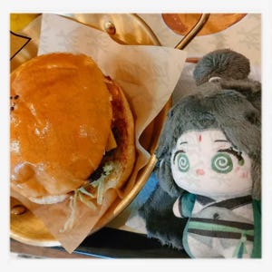 May include: A golden-brown burger with sesame seeds on top, served on a brown paper liner inside a gold-colored tray. A plush doll with green eyes and a green and white outfit is next to the burger.