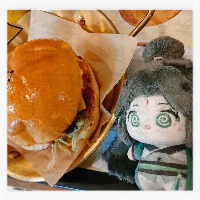 May include: A golden-brown burger with sesame seeds on top, served on a brown paper liner inside a gold-colored tray. A plush doll with green eyes and a green and white outfit is next to the burger.