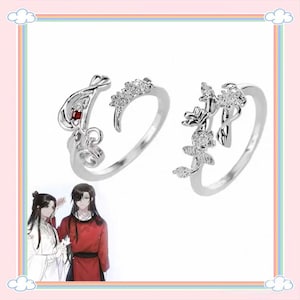 Puede incluir: Dos anillos plateados con diseños florales y abstractos. Un anillo presenta una piedra preciosa roja, el otro tiene detalles claros y brillantes. Los anillos están sobre un fondo blanco con un borde rosa y arcoíris.