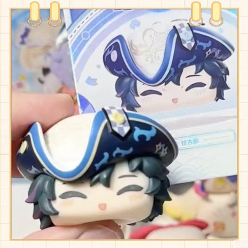 May include: A small, smiling figurine of a character wearing a blue and white hat with a decorative brim. The figurine has dark hair and is holding a matching card in the background. The hat has a yellow emblem.