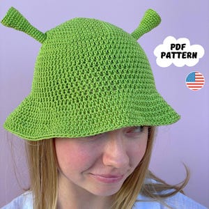 May include: A green crochet hat with two antennas, resembling a character from a popular movie. The hat is worn by a person with long blonde hair. The text "PDF PATTERN" is visible in a white cloud shape.
