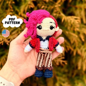 Op de afbeelding: Gehaakte amigurumi pop met roze haar, een rood jasje en gestreepte broek. De pop houdt een klein wit voorwerp in zijn hand. De pop wordt vastgehouden door een hand.